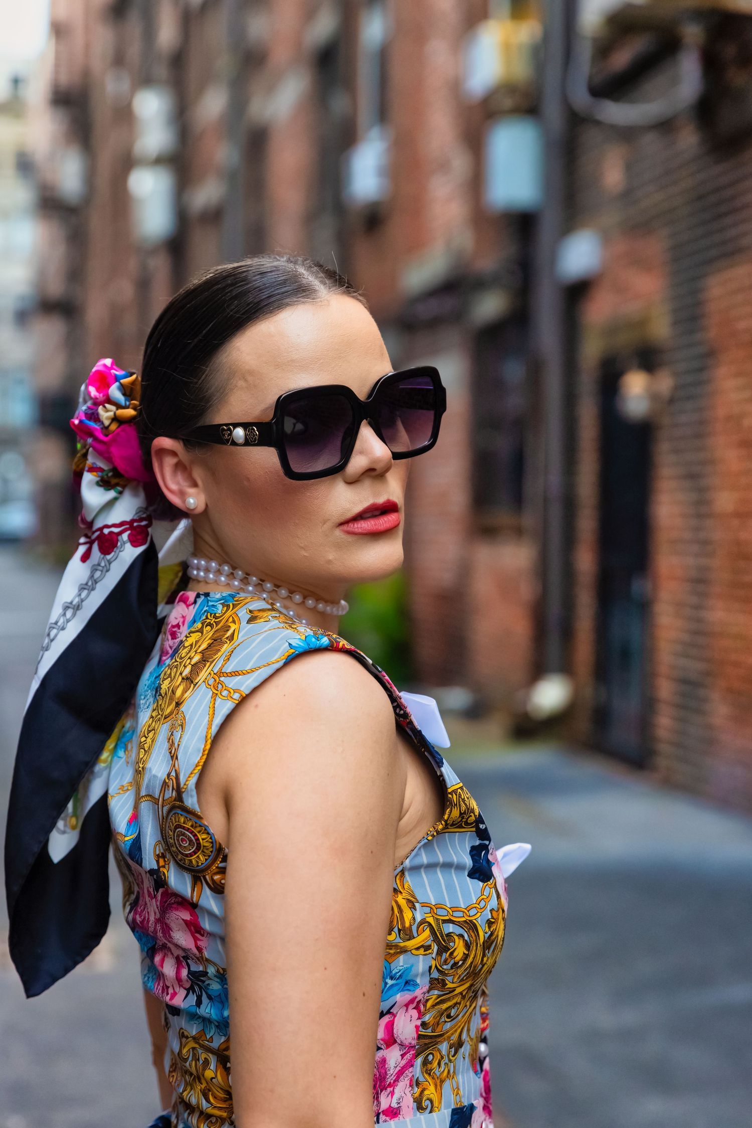 Stylish woman in floral dress, pearl necklace, silk scarf, and oversized sunglasses posing on a city alley street.