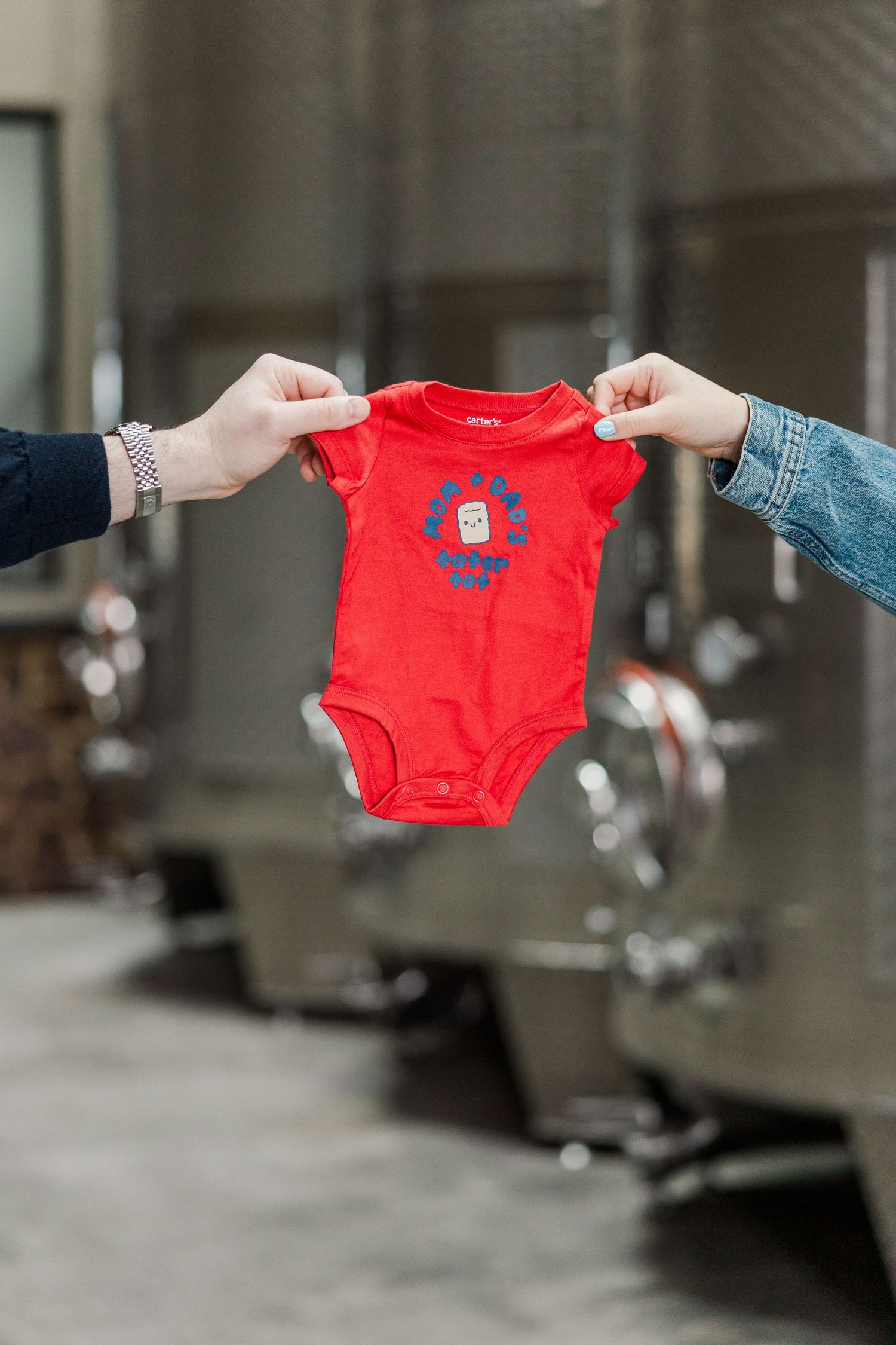 Two hands holding a red baby onesie with blue logo print in front of stainless steel wine tanks.