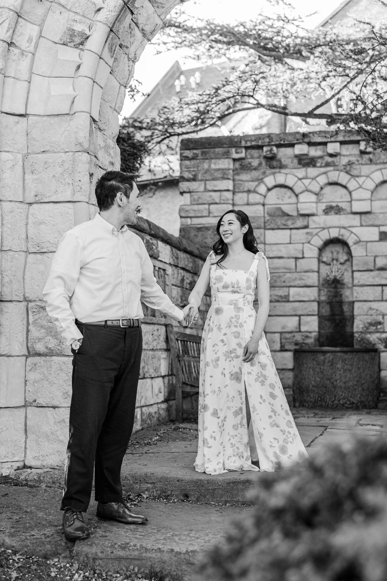 Black and white photo of a couple holding hands and smiling at each other in a stone garden setting.