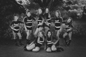 Eight young cheerleaders in matching dark uniforms pose together outdoors among shrubs in a black and white team portrait.
