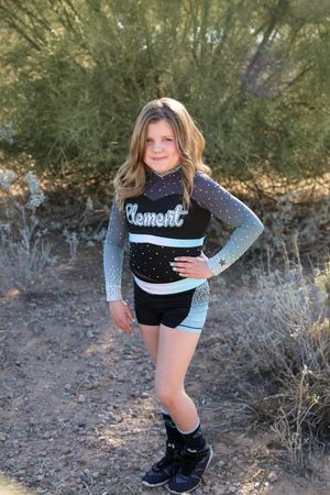 Girl with long wavy hair poses with a hand on her hip in a black and teal Element cheer uniform outdoors near desert shrubs.