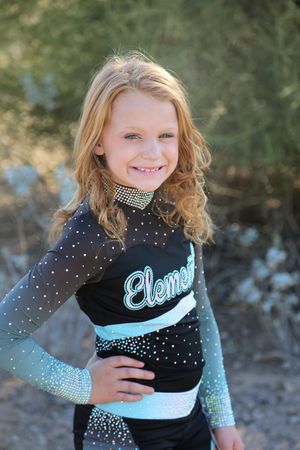 Smiling girl with wavy strawberry-blonde hair wears a black and teal rhinestone Element cheer uniform in front of green desert brush.