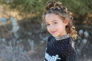 Young cheerleader with curly hair in a top bun smiles at the camera, wearing a rhinestone choker and a black Element cheer uniform.