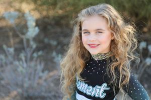 Smiling girl with long curly blonde hair wears a black and teal Element cheer uniform with a rhinestone choker near desert plants.