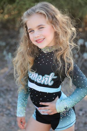 Cheerful girl with long curly blonde hair poses in a black and teal sparkly Element cheer uniform outdoors with a rhinestone choker.