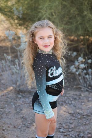 Smiling young girl with long curly blonde hair poses outdoors in a black and teal ombre cheer uniform with rhinestones.