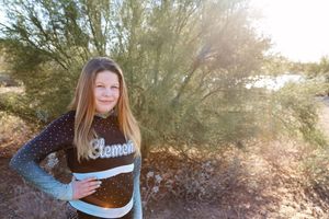 Girl with long straight hair smiles outdoors beside a desert shrub wearing a black and teal rhinestone cheer top.