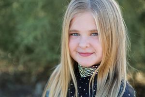 Close-up portrait of a young girl with long blonde hair and blue eyes wearing a rhinestone choker collar in a desert setting.