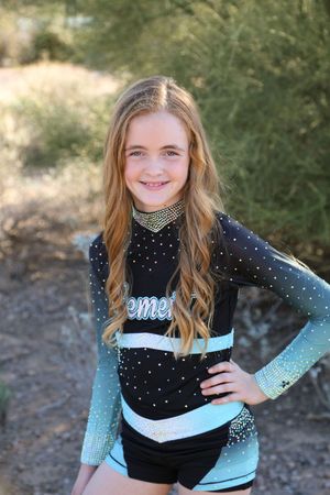 Happy girl with long wavy hair stands with hand on hip in a black and teal ombre cheer uniform with rhinestone details.