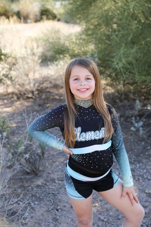 Smiling young cheerleader with long straight hair poses with hands on hips in a black and teal sparkly uniform outdoors.