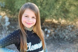 Young girl with long straight brown hair smiles in a black and teal Element cheer top with a rhinestone collar outdoors.