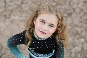 Smiling young girl with wavy blonde hair looks up at the camera wearing a sparkly black and teal cheer uniform outdoors.