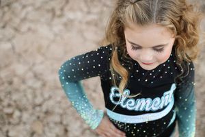Young girl looks down while posing outdoors in a black and teal ombre Element cheer uniform decorated with rhinestones.