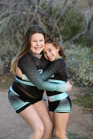 Two smiling young cheerleaders in black and teal sparkly uniforms hug each other outdoors in front of desert trees and shrubs.