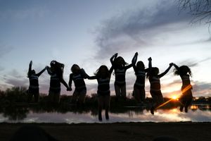Silhouettes of cheerleaders jumping with raised arms against a colorful sunset sky reflected over a calm lake.