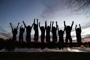 Silhouettes of a group of people jumping with arms raised in the air beside a lake under a purple and blue dusk sky.