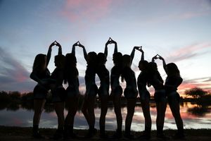 Silhouetted group of women standing in a line by a lake at sunset, raising their hands together to form heart shapes against a pink sky.