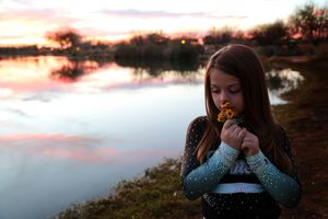 Girl in a sparkly cheer top holds yellow wildflowers to her face beside a lake with pink and purple sunset reflections.