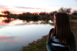 Girl in a cheer uniform gazes across a still lake at sunset, with soft pink clouds reflected in the water.