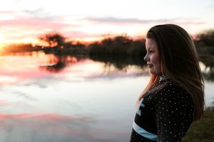 Profile of a girl with long hair looking toward the glowing sunset over a quiet lake in warm golden evening light.