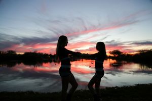 Two girls in silhouette hold hands and dance by a calm lake as a vivid pink and purple sunset reflects on the water.