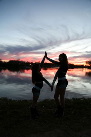Two cheerleaders in silhouette join raised hands beside a lake under a dramatic pink and blue sunset sky.