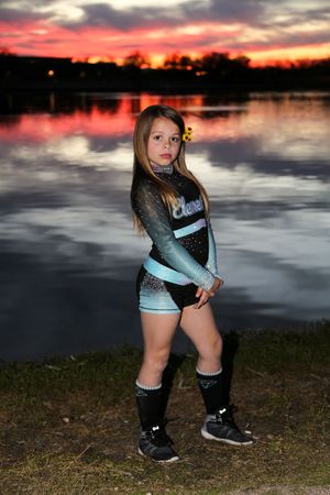 Girl in a teal and black cheer uniform with a yellow flower in her hair poses beside a lake at colorful dusk.