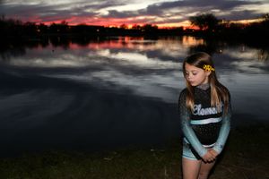 Young cheerleader with a yellow flower in her hair looks down by a dark lake under a red and gray sunset sky.
