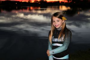Smiling girl in a cheer uniform with a yellow flower in her hair stands by a lake reflecting the evening sky.