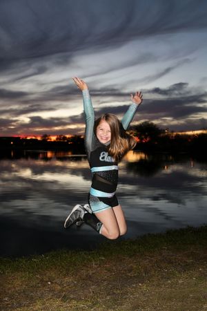 Young cheerleader in a teal and black uniform jumps joyfully with arms raised beside a lake under a dramatic sunset sky.