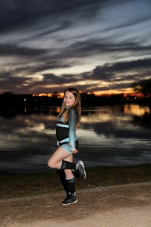 Young cheerleader in a sparkly teal and black uniform poses with one knee raised on a dirt path beside a sunset lake.