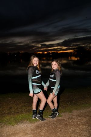 Two cheerleaders in matching teal and black uniforms hold hands and smile on the lakeshore as dusk settles.