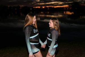 Two young cheerleaders in sparkly teal and black uniforms smile at each other while holding hands near a lake at twilight.