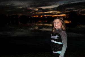Smiling girl in a black and teal cheer uniform poses outdoors by a dark lake with a glowing sunset behind her.