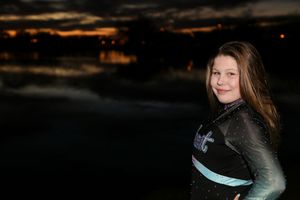 Girl with long brown hair in a sparkly cheer uniform smiles over her shoulder in front of a dark lake at sunset.