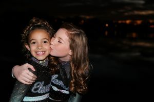 Teenage cheerleader kisses the cheek of a smiling younger girl, both wearing matching black and teal cheer uniforms at night.