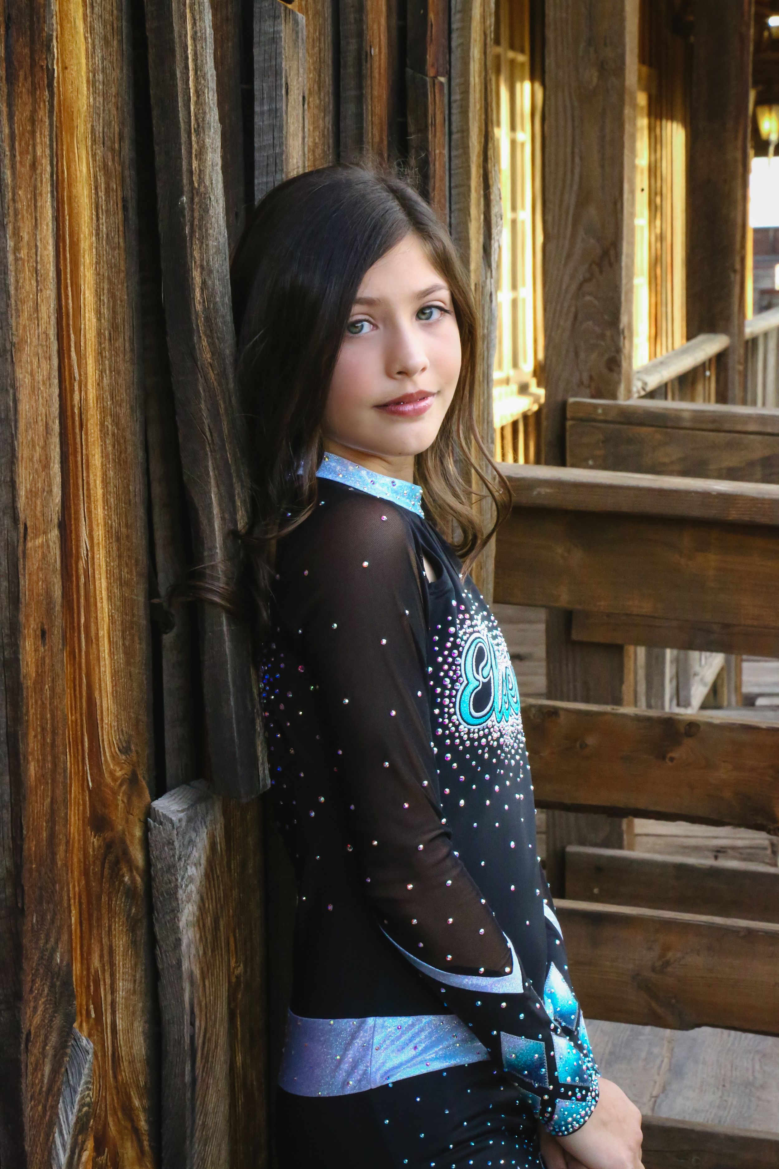 Young cheerleader in a black rhinestone uniform leans against a rustic wooden wall with a calm expression and blue eyes.