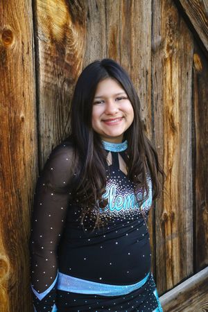 Smiling girl with long dark hair poses in a black and teal sparkling cheer uniform beside a weathered wooden wall.