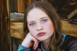 Close-up portrait of a young girl with blue eyes resting her chin on her hand in front of weathered wooden planks.