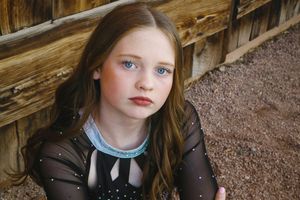 Girl with long wavy hair and blue eyes sits on gravel beside a rustic wooden wall wearing a black cheer uniform with a teal collar.