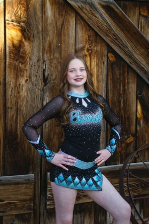 Smiling cheerleader in a black and teal Elements uniform with sheer sparkly sleeves poses with hands on hips by an old wooden barn wall.
