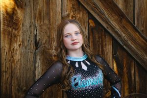 Young cheerleader with long wavy hair wears a black and teal Elements uniform and poses in front of a rustic wooden barn door.