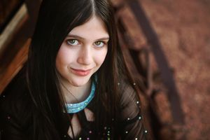 Smiling girl with long dark hair and light eyes wears a teal collar and a black sparkly top while looking up at the camera.