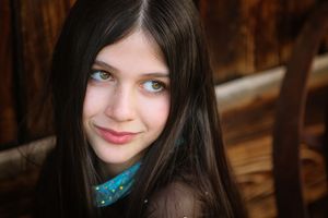 Portrait of a girl with long straight dark hair and green eyes looking off to the side with a soft smile near rustic wood.