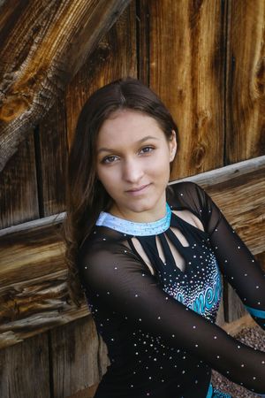 Teen cheerleader with brown hair wears a black sparkly uniform with a light blue collar and poses against weathered wooden planks.