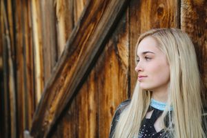 Blonde young woman with long hair gazes to the side while standing beside a weathered rustic wooden barn wall.