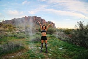 Woman in athletic wear and knee-high socks raises her arms joyfully amid drifting powder in a green desert landscape with rocky cliffs.