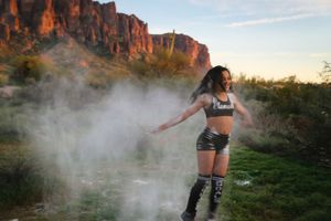 Dancer in a crop top and shorts spins with outstretched arms through a cloud of powder at sunset below red desert cliffs.