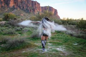 Motion blur captures a dancer twirling with swirling white powder against a backdrop of desert brush and red rock cliffs.