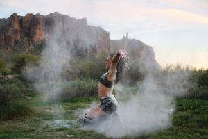 Woman kneels on the grass with arms raised as clouds of white powder swirl around her beneath a rugged desert mountain.